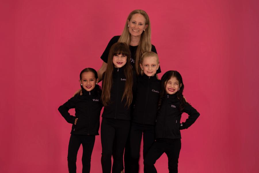 Celebrate Recital Month, A smiling dance instructor stands behind four young dancers in matching black outfits with their names embroidered, posing in front of a bright pink background.